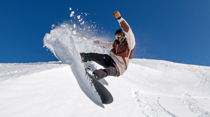 Australian Paralympian snowboarder Ben Tudhope, kicking up snow mid-air, while snowboarding down a mountain.