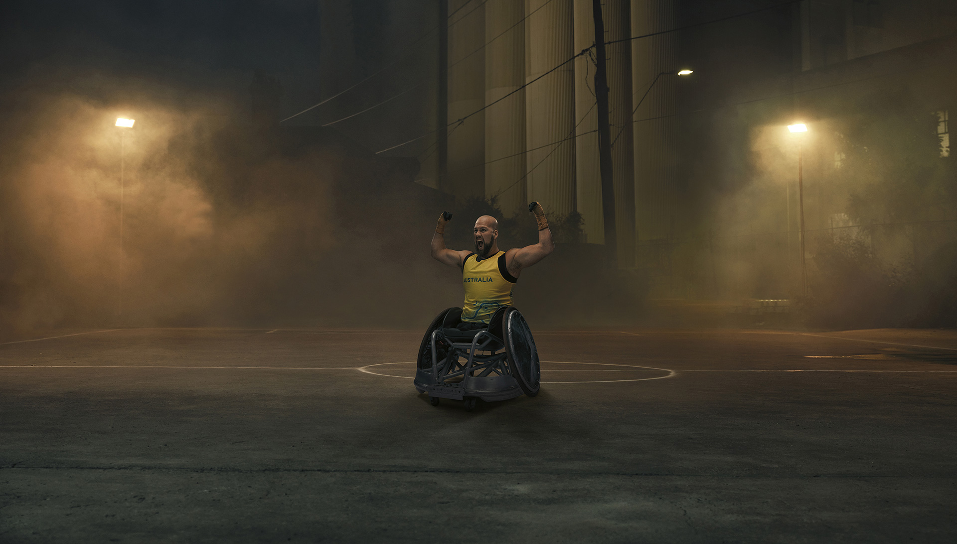 Australian Paralympian wheelchair rugby player Ryley Batt, posing with his arms flexed upwards, on a moodily lit, foggy basketball court at night.