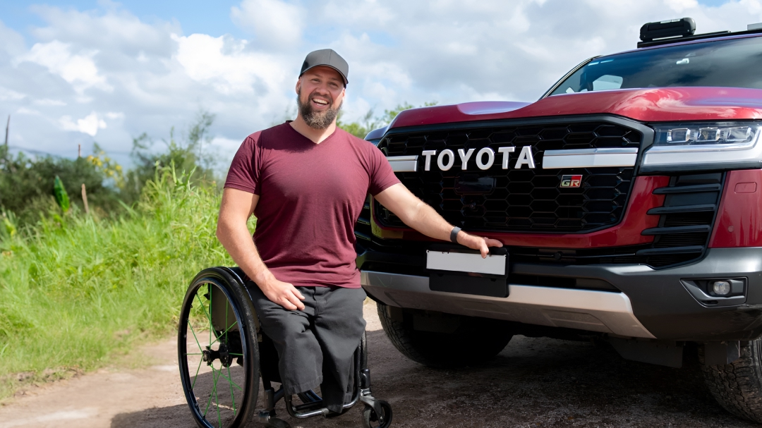 Australian Paralympian wheelchair rugby player Ryley Batt proudly smiling in front of his red Toyota LandCruiser 300, next to a grassy field.