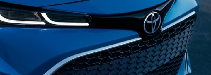 An artistically photographed front grille and left headlight of a blue 2024 Toyota Corolla in the ZR grade, headlights on, in a cool, moody atmosphere. 