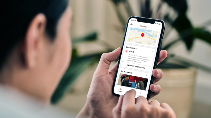 A woman with a smartphone displaying the myToyota Connect app. The screen shows a map, information about safety recalls, and an option to book a service.
