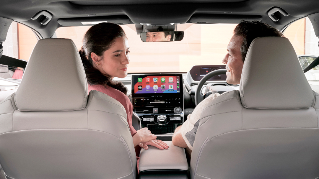 A man and woman in the front seats of a Toyota, smiling, with the woman in the passenger seat turning her head looking towards the back passenger seat behind the driver.