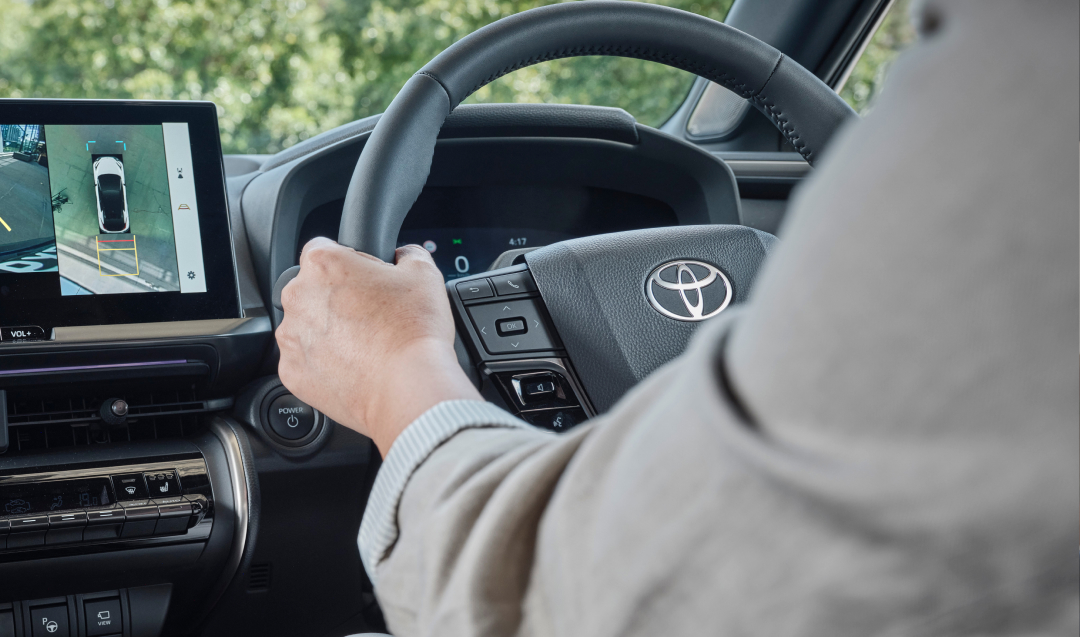 A man in a gray suit driving a Toyota. The digital information display next to the steering wheel shows a top-down view of the car and its surroundings to assist with parking.