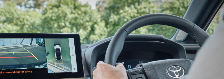 A man in a gray suit driving a Toyota. The digital information display next to the steering wheel shows a top-down view of the car and its surroundings to assist with parking.