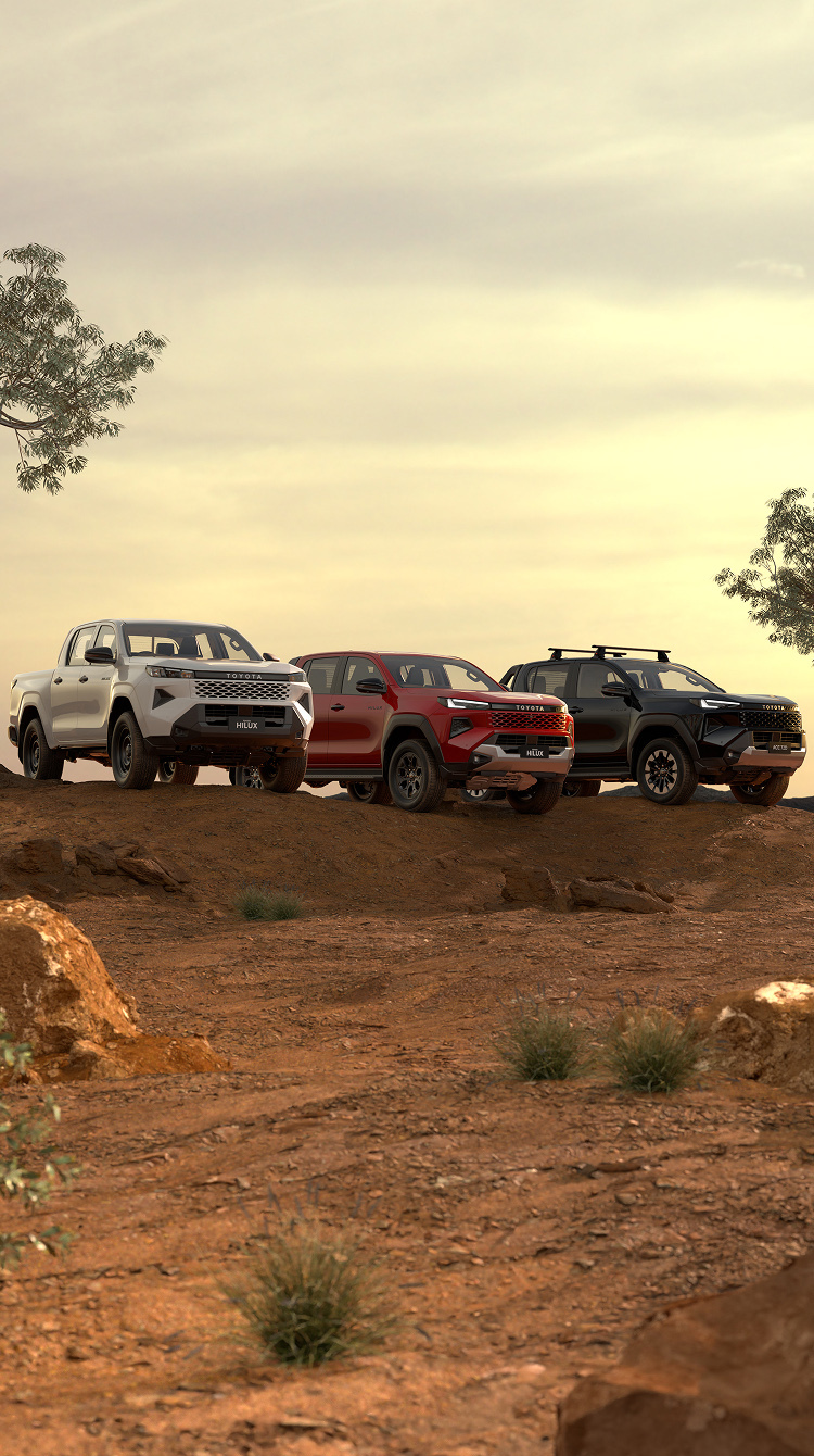 Three HiLux double-cab utes sit parked in a dirt clearing at golden hour. The first has Glacier White paint, the middle one is Feverish Red and the end one is Ash Slate. Australian trees and scrub frame the scene, while the sun sets behind them. 