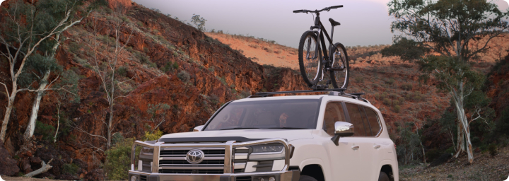 A sandy taupe coloured LandCruiser 70 with hay bails in the tray pulls a grey caravan along a red dirt track. 