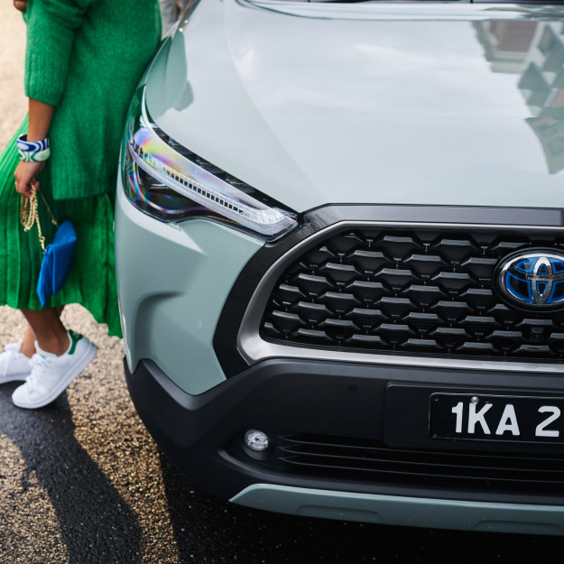 A woman in a green dress and white shoes leaning against the side of a khaki Corolla Cross.
