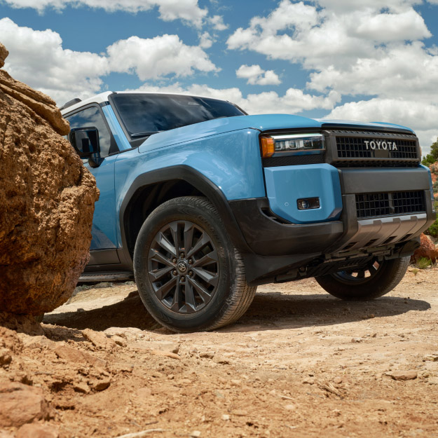 A blue Prado Altitude driving in an arid environment past a large orange rocky boulder in the middle of the day.