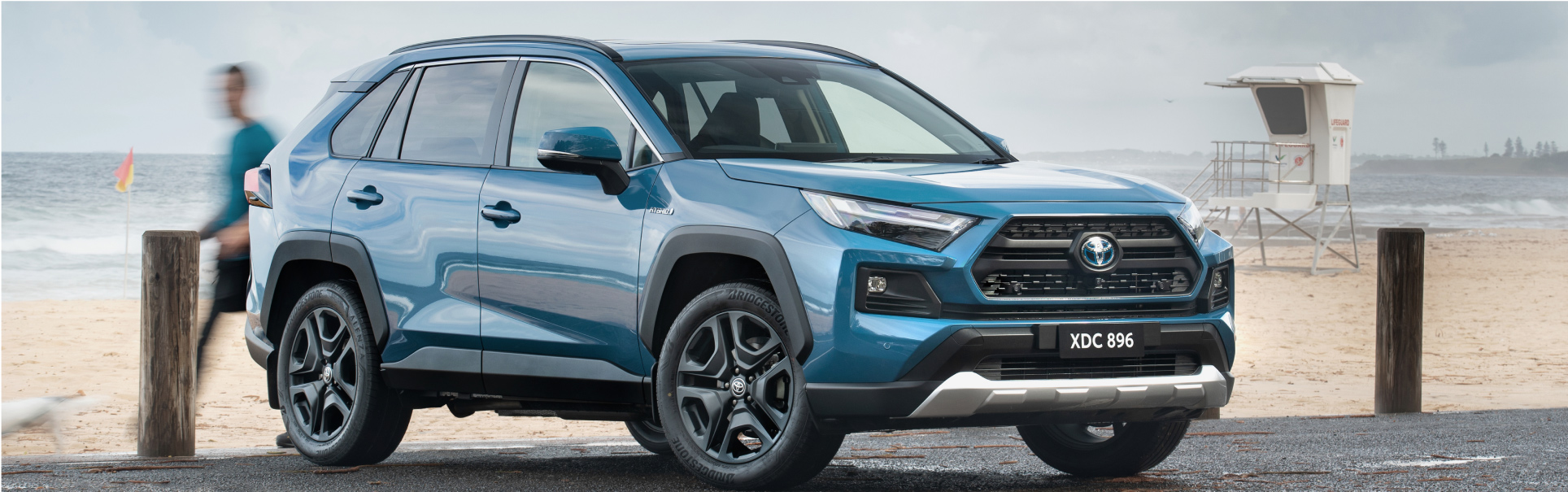 A blue RAV4 Edge parked alongside a beach, with people walking by, stormclouds rolling in.