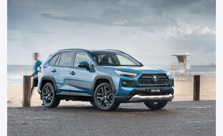 A blue RAV4 Edge parked alongside a beach, with people walking by, stormclouds rolling in.