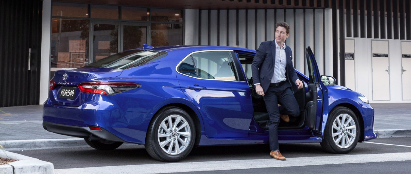 A man in a suit getting out of a Blue Toyota Camry on a modern city street.