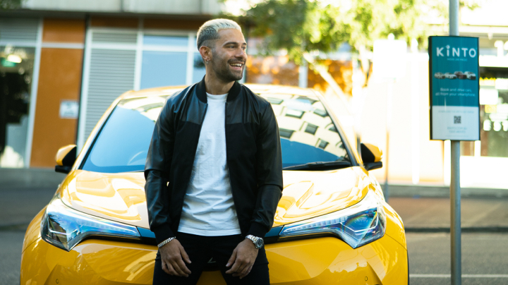 A man resting against the bonnet of a yellow car, grinning happily on a sunny afternoon.
