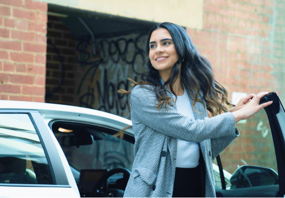 A woman getting out of the driver’s side of a white car, a smile on her face as her hair flows in the breeze.