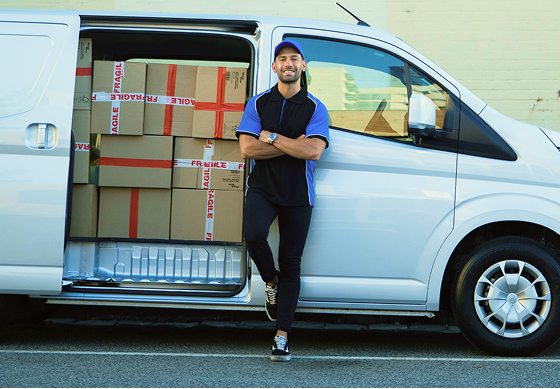 A man standing confidently, arms crossed and resting against a white delivery van, smiling. Its door is open, showing the stacks of boxes inside.