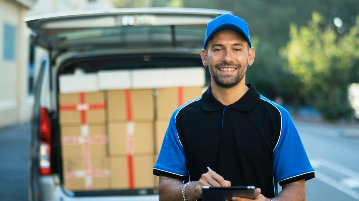 A man in a blue delivery uniform, holding a digital tablet and smiling, a loaded delivery van in the background.