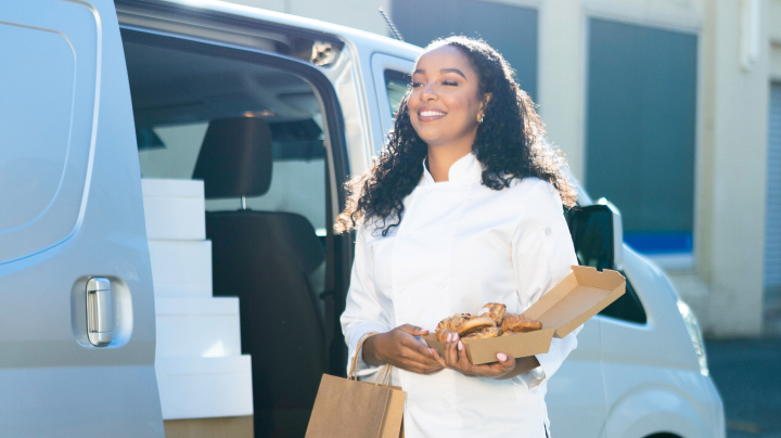 A woman in a chef’s uniform carrying baked goods from a white delivery van, sun shining down on her as she smiles.